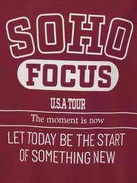 Vinröd t-shirt med vit fet text: "SOHO FOCUS U.S.A TOUR" och "LÅT IDAG VARA STARTEN PÅ NÅGOT NYTT," i olika teckenstorlekar.