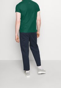 Un homme portant une chemise à manches courtes vert foncé, un pantalon bleu marine, des chaussettes blanches et des chaussures légères gris clair, se tenant face à un mur blanc.