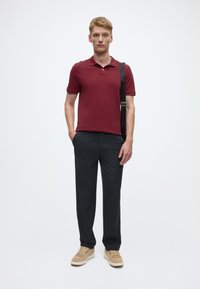Red polo shirt, dark tailored pants, and beige sneakers. Standing pose with one hand in pocket, neutral background. Simple and modern design.