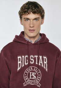 Bordeauxroter Hoodie mit einem Frontdruck, der "BIG STAR" und ein rundes Logo mit "YOUR LIFE SINCE 1979" zeigt. Er hat eine Kapuze mit Kordelzug.