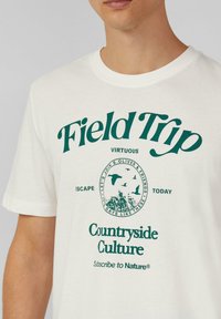 Weißes Baumwoll-T-Shirt mit grüner Schrift und kreisförmigem Grafik. Das Design beinhaltet von der Natur inspirierte Elemente und Slogans, die mit der Kultur des Landlebens in Verbindung stehen.