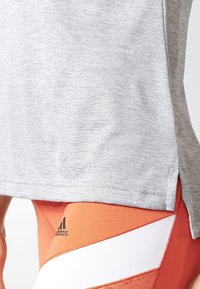 Harmaa urheilupaita, jossa on teksturoitu pinta ja sivuventtiilit, yhdistettynä oransseihin legginsseihin, joissa on musta logo ja valkoinen raidallinen yksityiskohta.