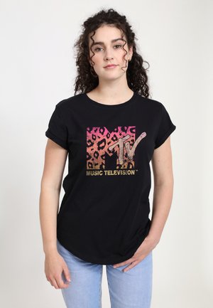 Vrouw met krullend donker haar, gekleed in een zwart MTV Music Television T-shirt met roze en oranje luipaardprint, gecombineerd met lichtblauwe jeans.