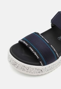 Sandale noire avec semelle blanche mouchetée, brides élastiques bleu marine et texte blanc indiquant "See by Chloe" sur les brides.