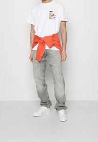 Witte katoenen t-shirt met een meerkleurige bedrukte zak, lichtgrijze spijkerbroek en witte sneakers; oranje sweatshirt om de taille geknoopt.