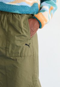 Olivenfargede cargo-bukser med sidelommer, med svart Puma-logo. Brukt sammen med en flerfarget, strukturert fleecegenser.