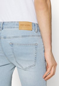 Vaqueros de mezclilla azul claro con una etiqueta de cuero en la cinturilla con el texto "ONLY & SONS". Los vaqueros tienen bolsillos traseros con detalles de costura.