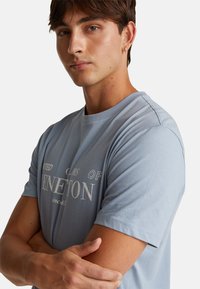 Mladý muž s krátkými hnědými vlasy, oblečený v světle modrém tričku Benetton, se zkříženýma rukama a malým kruhovým náušnicí v levém uchu.