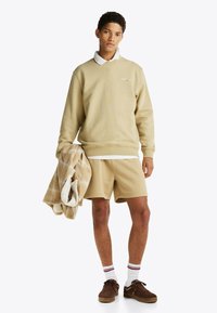 Sudadera beige con puños acanalados, superpuesta sobre una camisa blanca con cuello. Pantalones cortos beige y una manta de cuadros. Zapatillas marrones y calcetines a rayas.