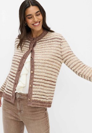 Frau lächelt mit geschlossenen Augen, trägt eine braun-creme gestreifte Strickjacke über einer weißen, gerafften Bluse und braune Hosen.