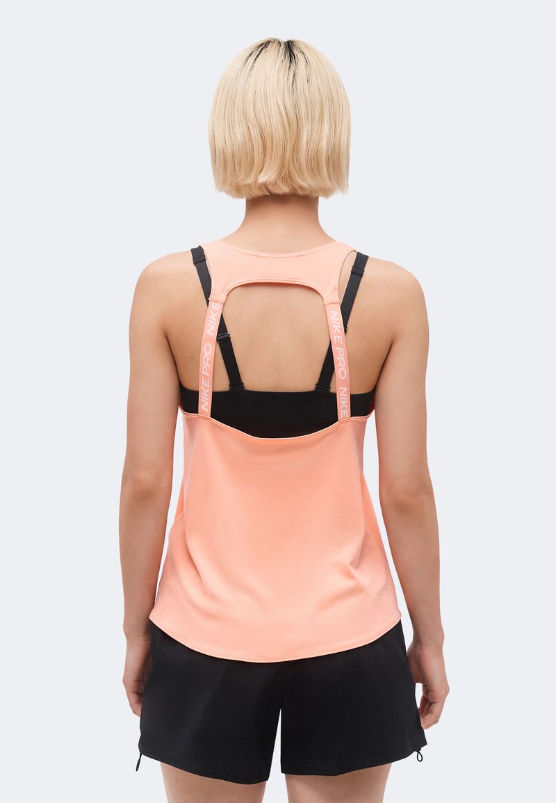 Άτομο με κοντά ξανθά μαλλιά που φοράει ένα ροδακινί τοπ με ανοιχτή πλάτη πάνω από μαύρο αθλητικό σουτιέν και μαύρα σορτς, με την πλάτη γυρισμένη προς την κάμερα.