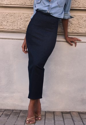 Persona que lleva una falda ajustada de denim oscuro, una camisa de denim azul claro con las mangas remangadas y sandalias doradas con tiras, apoyada contra una pared con textura.