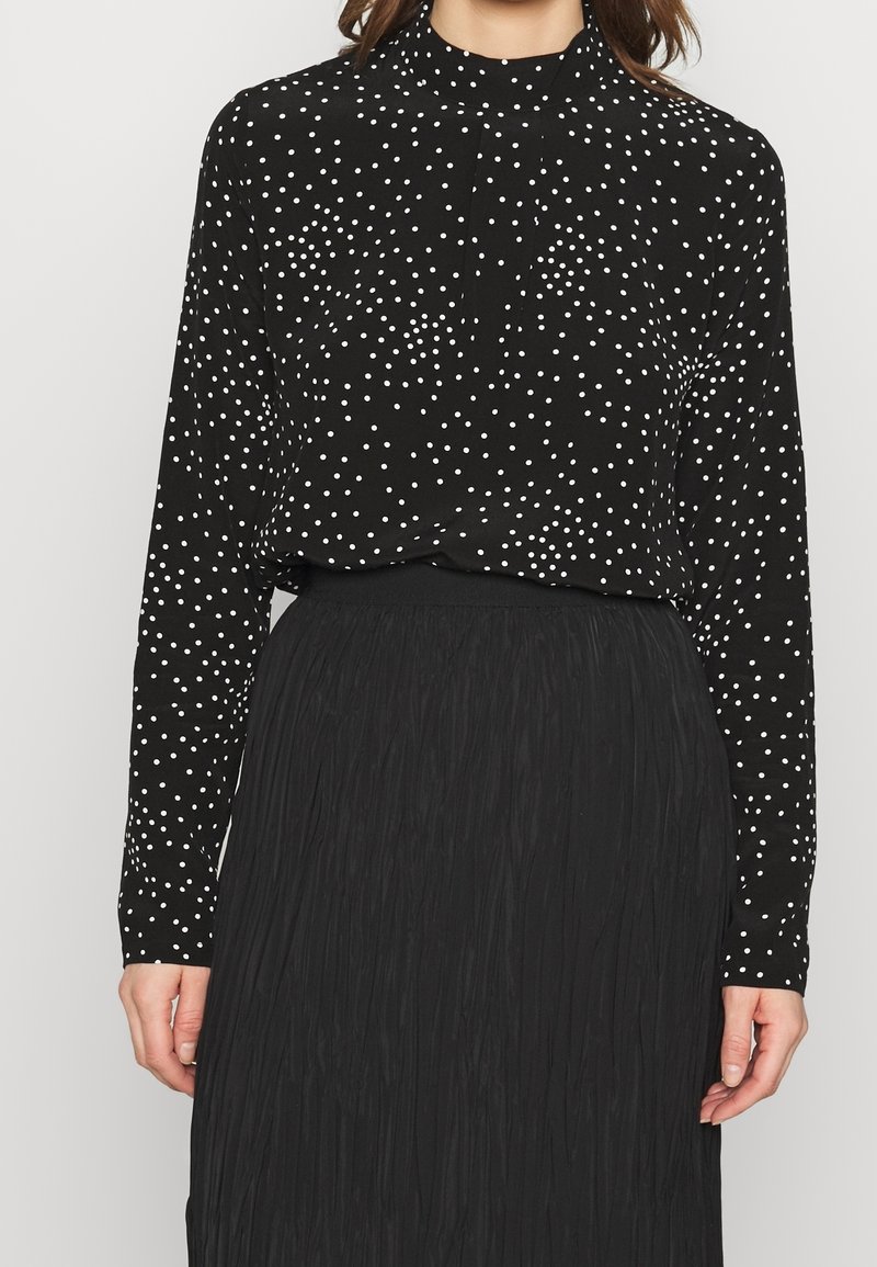 Femme portant un haut noir à manches longues avec des pois blancs, rentré dans une jupe noire texturée taille haute, se tenant devant un fond blanc.