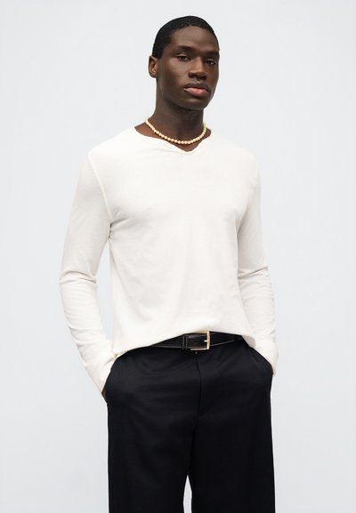 Άνδρας με άσπρο πουκάμισο με μακριά μανίκια, μαργαριταρένιο κολιέ, μαύρο παντελόνι και μαύρη ζώνη, με τα χέρια στις τσέπες, μπροστά σε απλό φόντο.