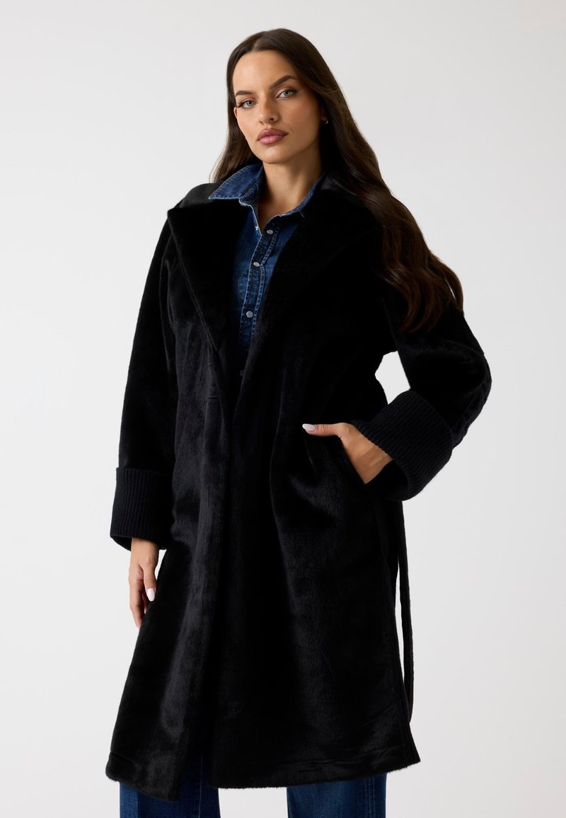 Schwarzer Pelzmantel mit weitem Kragen, tailliertem Gürtel und gerippten Ärmelbündchen. Weiche Textur und knielanger Schnitt. Taschen auf beiden Seiten.