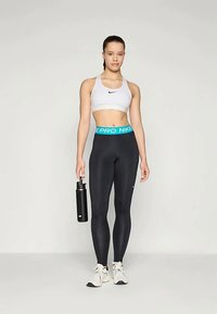 Fehér sportmelltartó fekete Nike logóval, magas derekú fekete leggings kék Nike Pro derékkal, vízpalackot tartva, fehér sportcipőt viselve.