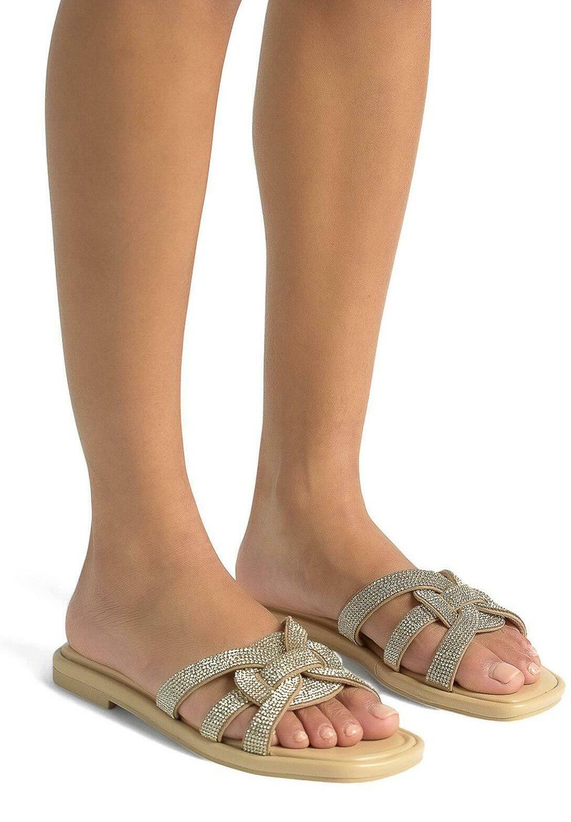Beige platte slidesandalen met een geweven ontwerp en een accent van rhinestones. De zool is plat en de banden zijn breed, waardoor de tenen blootgesteld worden.