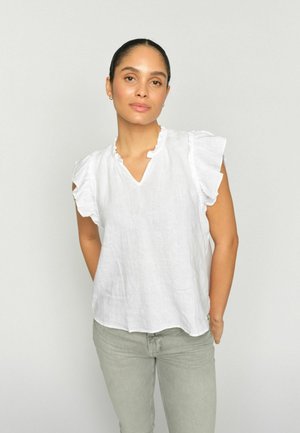 Žena so zviazanými vlasmi, oblečená v bielej blúzke bez rukávov s volánmi a svetlosivých nohaviciach, stojaca pred jednoduchým svetlým pozadím.