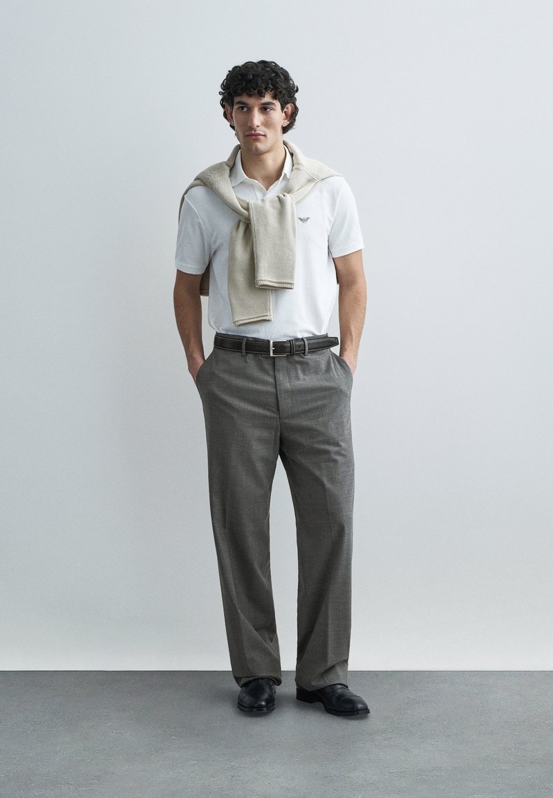 Weißes Poloshirt, graue gemusterte Hose, beiger Pullover lässig über die Schultern gelegt, schwarzer Gürtel und schwarze Schuhe vor neutralem Hintergrund.