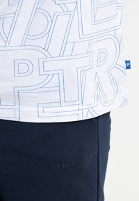 Camiseta blanca con diseño de texto contornado en azul, combinada con pantalones de chándal marinos que presentan un logotipo de marca en relieve. Material de algodón con textura suave.