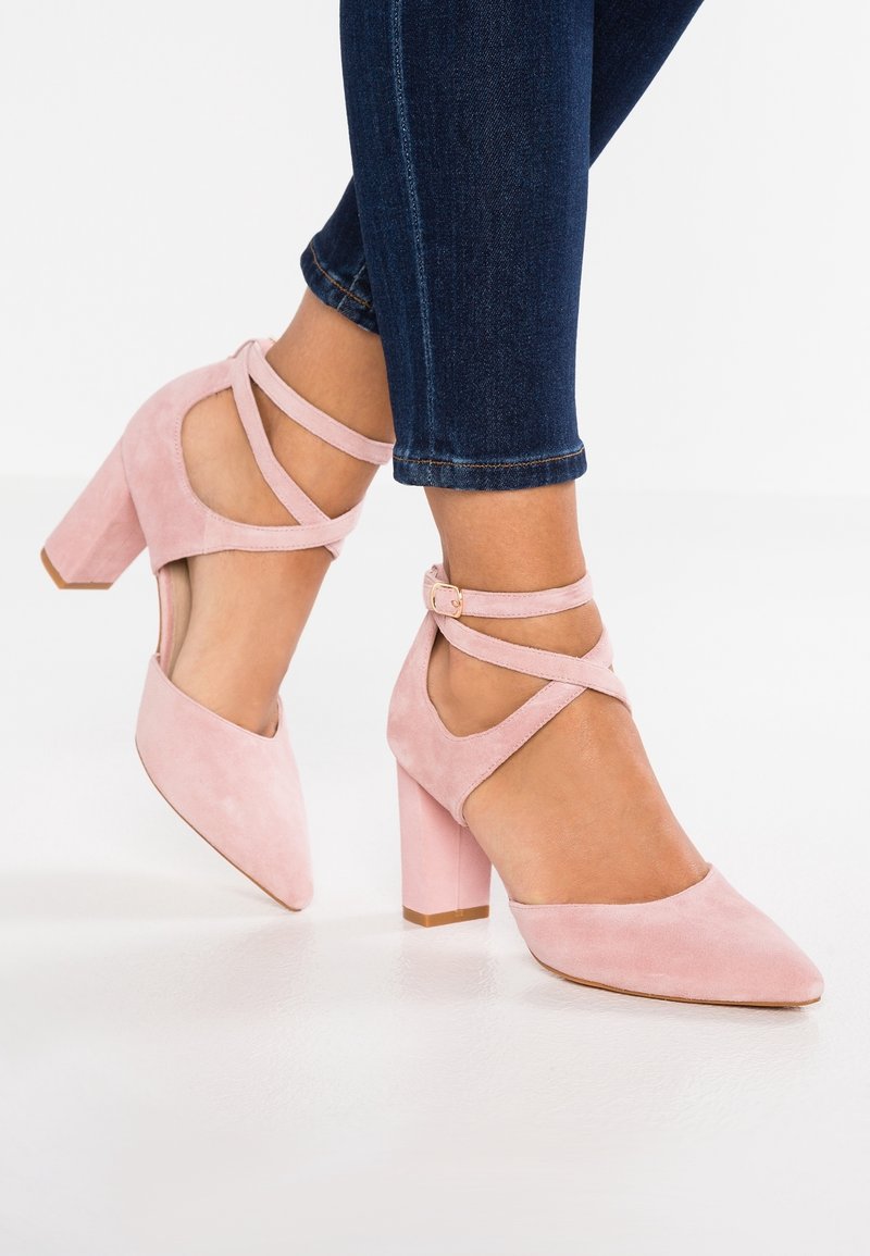 Femme portant des chaussures à talons carrés en daim rose avec des brides croisées autour de la cheville, associées à un jean skinny bleu foncé.