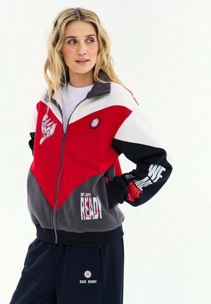 Blondynka ubrana w kurtkę z kolorowymi blokami w kolorach czerwonym, białym, czarnym i szarym, zapinaną na zamek, z naszywkami Bugs Bunny, oraz czarne spodnie z napisem Bugs Bunny.
