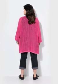 Cardigan lavorato a maglia oversize di un vivace colore rosa, con una vestibilità comoda e texture a coste, abbinato a jeans neri cropped e scarpe nere.