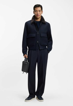 Giacca bomber blu navy con colletto in pelliccia sintetica, due tasche frontali, abbinata a pantaloni scuri e sneakers blu navy, con in mano una piccola borsa nera.