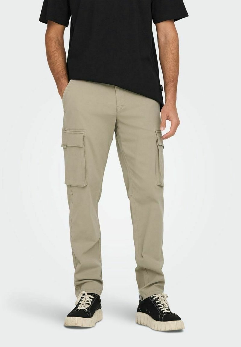 Homme portant un pantalon cargo beige, des baskets noires avec semelles blanches et un t-shirt noir, debout avec une main dans la poche.