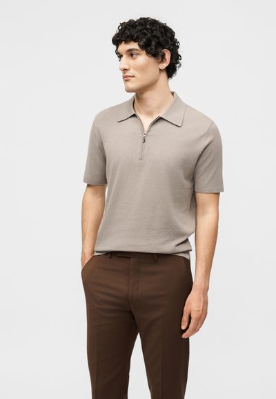 Homme aux cheveux bouclés portant une chemise beige à manches courtes avec col zippé et un pantalon marron, debout les mains dans les poches, regardant sur le côté.