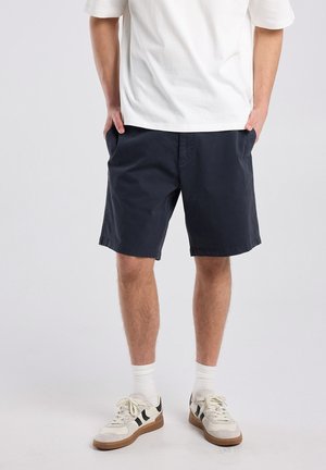 Man die een wit T-shirt, marineblauwe korte broek, witte sportsokken en witte sneakers met zwarte strepen draagt, met handen in de zakken staat.