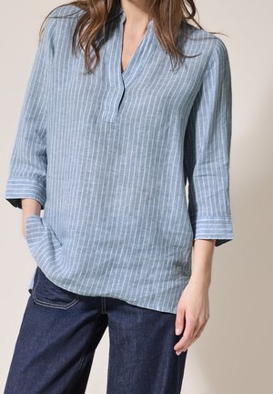 Kvinde iført en lyseblå bluse med vertikale striber og trekvartlange ærmer samt mørkeblå jeans, hånd i lommen.
