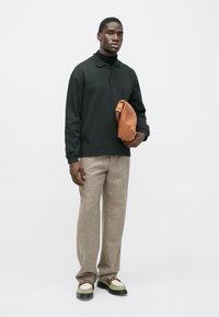 Čierna polokošeľa s dlhým rukávom, sivé tvídové nohavice, zeleno-biele topánky. Drží hnedú koženú tašku. Jednoduchý dizajn a textúrované materiály.