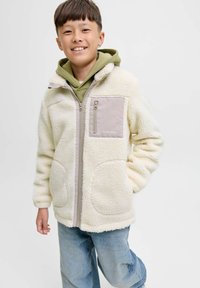 Crème-kleurige fleecejas met een rits aan de voorkant, met een grijze borstzak en twee zijzakken. Draag het over een groene hoodie.
