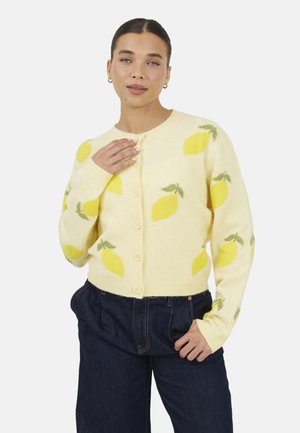 Vrouw met een geel vest met knopen en citroenpatronen en donkerblauwe hoge taillebroek, poserend met één hand op de borst en de andere in de zak.