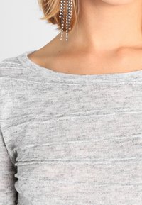 Szary sweter z miękkim, teksturowanym splotem, w poziome paski i szerokim dekoltem, uzupełniony błyszczącymi kolczykami wiszącymi.