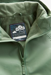 Intérieur d'une veste verte imperméable avec un doux doublure en polaire, présentant une fermeture éclair et une étiquette colorée affichant "RAIN WEAR" ainsi que des détails de design.