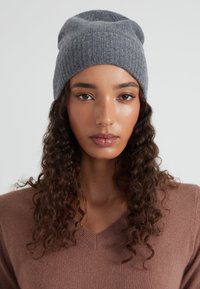Gorro de punto gris con diseño acanalado texturizado, ajuste ligeramente caído, combinado con un suéter marrón de cuello en v en un entorno neutral.