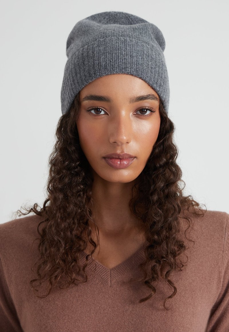 Gorro de punto gris con diseño acanalado texturizado, ajuste ligeramente caído, combinado con un suéter marrón de cuello en v en un entorno neutral.