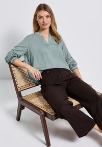 Blouse verte clair à manches longues avec un col en V, associée à un pantalon noir évasé, assise sur une chaise en bois avec un siège en osier.