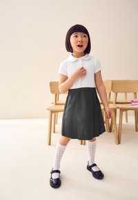 Fehér, rövid ujjú ing, kerek gallérral, amelyet egy sötét redőzött szoknyával párosítottak, fehér mintás, térd fölé érő zoknival és fekete cipővel.