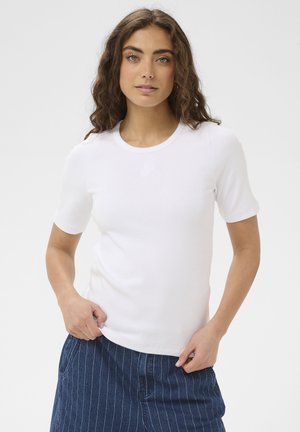 Femme aux cheveux bouclés portant un t-shirt blanc uni à manches courtes et un pantalon bleu à fines rayures, debout devant un fond blanc.