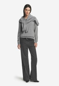 Frau trägt einen grauen Pullover mit einem weiteren über den Schultern gebunden, dunkle hochtaillierte weite Jeans, schwarzen Gürtel und schwarze spitze Schuhe.
