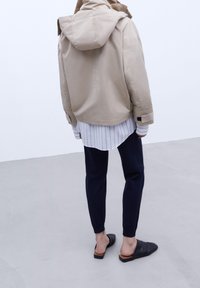 Persona con una giacca a vento beige con cappuccio, una maglietta a righe, pantaloni jogger neri e scarpe slip-on nere in piedi su un pavimento grigio contro un muro bianco.
