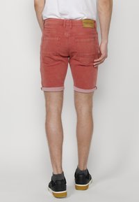 Pantalones cortos de mezclilla roja con dobladillo vuelto, mostrando bolsillos traseros y una etiqueta de cuero de la marca en la cintura. Combinados con zapatillas negras.