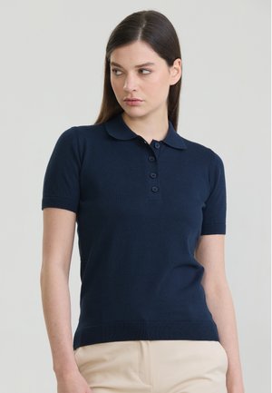 Kvinde med glat brunt hår iført en marineblå kortærmet polo og beige bukser, der ser let mod venstre.