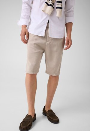 Homme portant un short beige jusqu'aux genoux, une chemise blanche boutonnée, un pull rayé drapé sur les épaules, et des mocassins marron foncé.