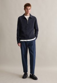 Granatowy sweter z zamkiem na wysokości szyi, wykonany z fakturowanej tkaniny, noszony na białej koszuli. Połączony z luźnymi dżinsami i czarnymi loafersami.