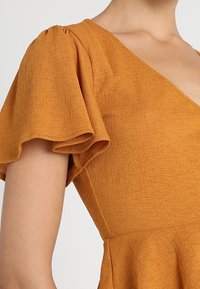 Hořčičkově žluté šaty s balónovými rukávy, strukturovaná látka a výstřih do V. Hrudník má peplum lem, který zvýrazňuje siluetu.