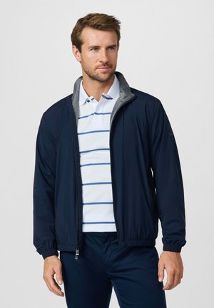 Mann trägt eine marineblaue Zip-up-Jacke über einem weißen Poloshirt mit blauen Streifen, kombiniert mit marineblauen Hosen, vor einem schlichten Hintergrund stehend.
